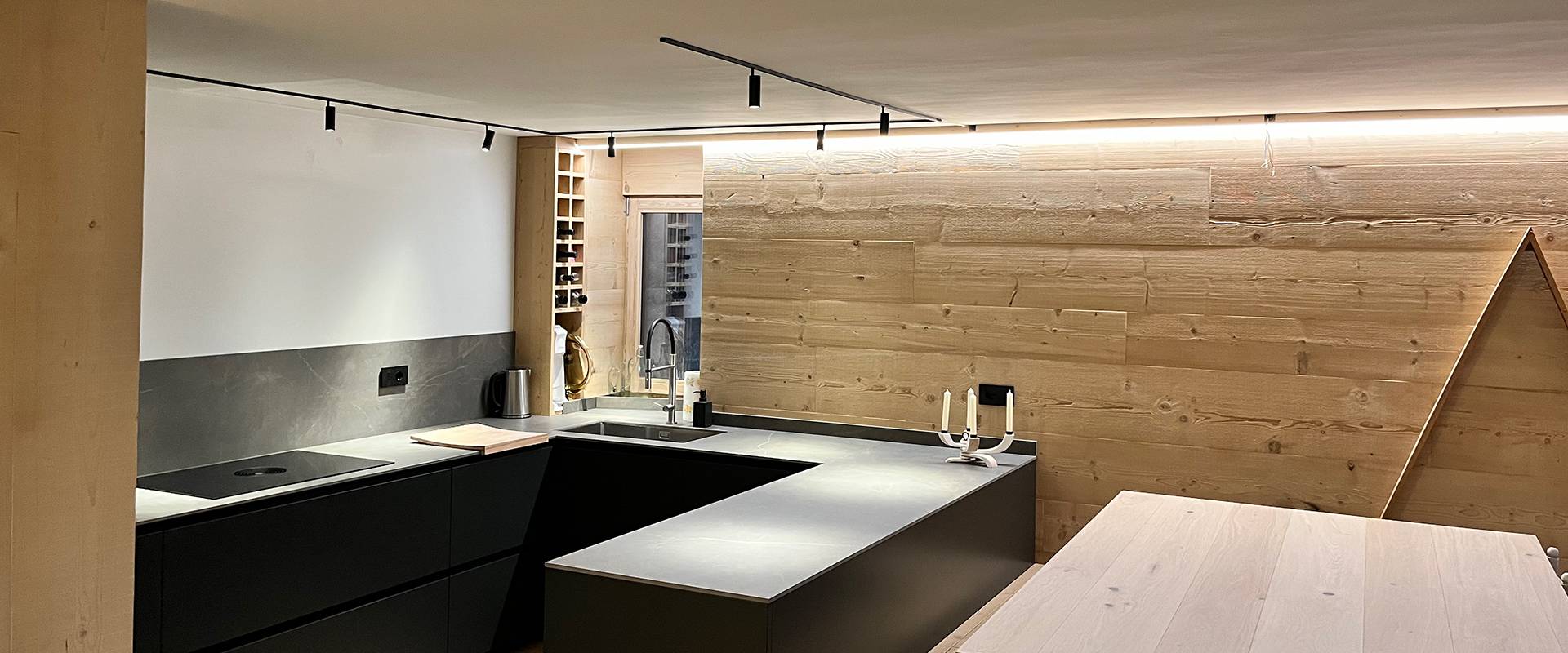 Cucina open space moderna con isola centrale e arredi in legno FGV Impianti Biella S.R.L. Cucina open space moderna con isola centrale e arredi in legno. Interno di una cucina ad induzione open space dal design contemporaneo. Da un lato troviamo un atgliere di legno, un bollitore in acciaio, un mobile porta bottiglie, una finestra e il lavandino. Al centro si trova un’isola scura con piano di lavoro su cui è appoggiato un candelabro moderno bianco a 4 bracci. e un tavolo in legno chiaro. Le pareti sono rivestite in pannelli lignei e i mobili hanno finiture nere opache. Il soffitto presenta una striscia luminosa a led che corre lungo il bordo e un binario con faretti orientabili. Sul lato destro pensile a forma triangolare