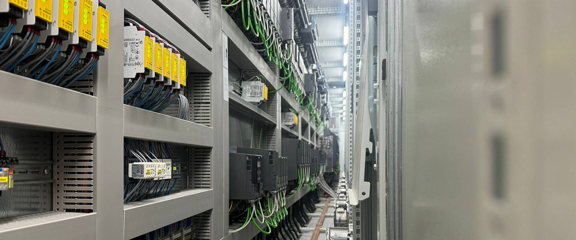 Quadro elettrico industriale con dispositivi e cablaggi ordinati FGV Impianti Biella S.R.L. Armadio elettrico industriale visto in prospettiva. Al suo interno ci sono file di dispositivi elettronici, moduli di controllo, PLC e relè montati su rack. I cavi verdi e neri scorrono ordinati nelle canaline, con etichette di identificazione. Il corridoio tecnico si estende in profondità mostrando altri quadri allineati lungo la parete.