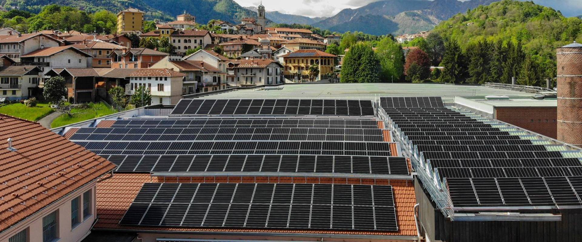 Pannelli solari su tetti di edifici con paesaggio montano FGV Impianti Biella S.R.L. Pannelli solari su tetti di edifici con paesaggio montano Ampia superficie di pannelli fotovoltaici neri installati sui tetti rossi di un complesso industriale. In secondo piano si vedono altre case del paese circondate da alberi. Sullo sfondo si ergono montagne verdi e boscose. Il cielo sopra è parzialmente coperto da nuvole bianche e grigie, con tratti di azzurro visibile.