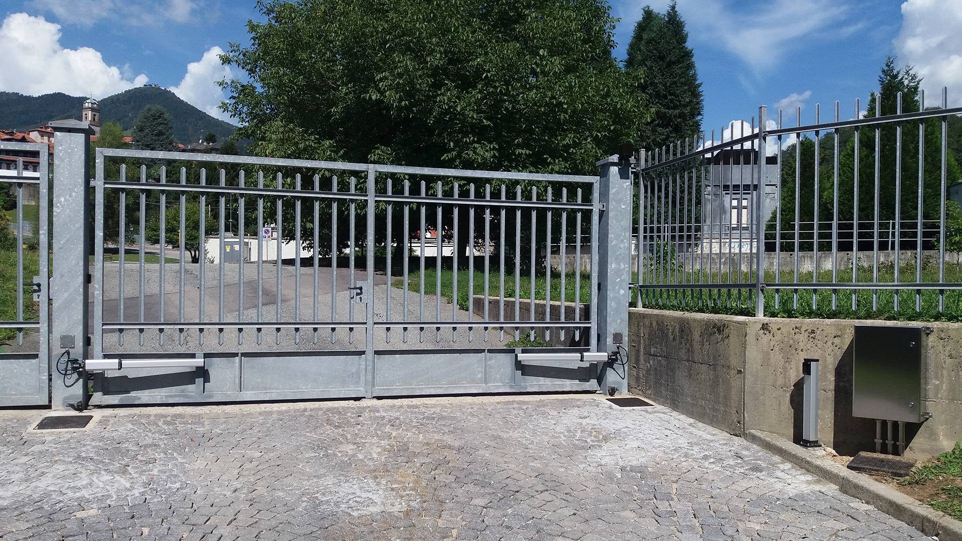 Cancello scorrevole metallico automatizzato con paesaggio montano FGV Impianti Biella S.R.L. Cancello scorrevole in metallo zincato montato su un vialetto pavimentato in pietra. Alla base, sul lato destro, è visibile il motore per l’apertura automatizzata. Il cancello è affiancato da colonne e ringhiere dello stesso materiale. Sullo sfondo si trovano un prato, diversi alberi verdi, case sparse e montagne coperte di vegetazione, sotto un cielo azzurro con nuvole bianche.