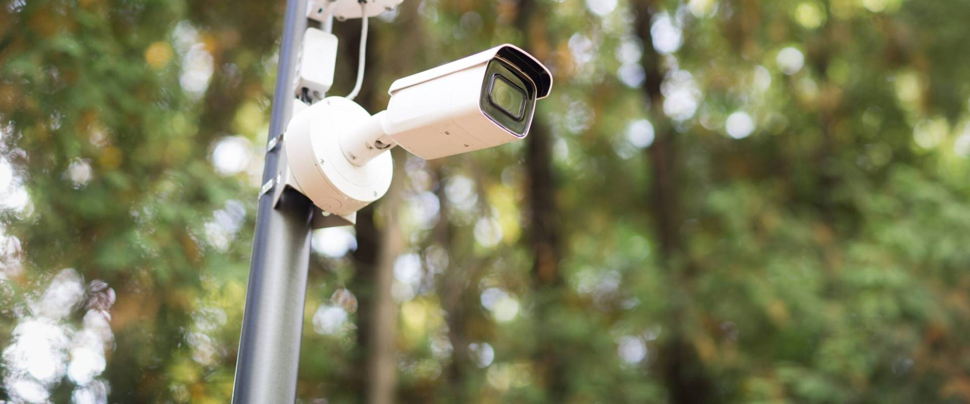 Telecamera di sicurezza bianca montata su supporto esterno FGV Impianti Biella S.R.L. Telecamera di sorveglianza bianca di tipo bullet, fissata a un supporto metallico esterno. L’obiettivo è orientato frontalmente. Sullo sfondo si distinguono alberi verdi sfocati illuminati dalla luce naturale del giorno.