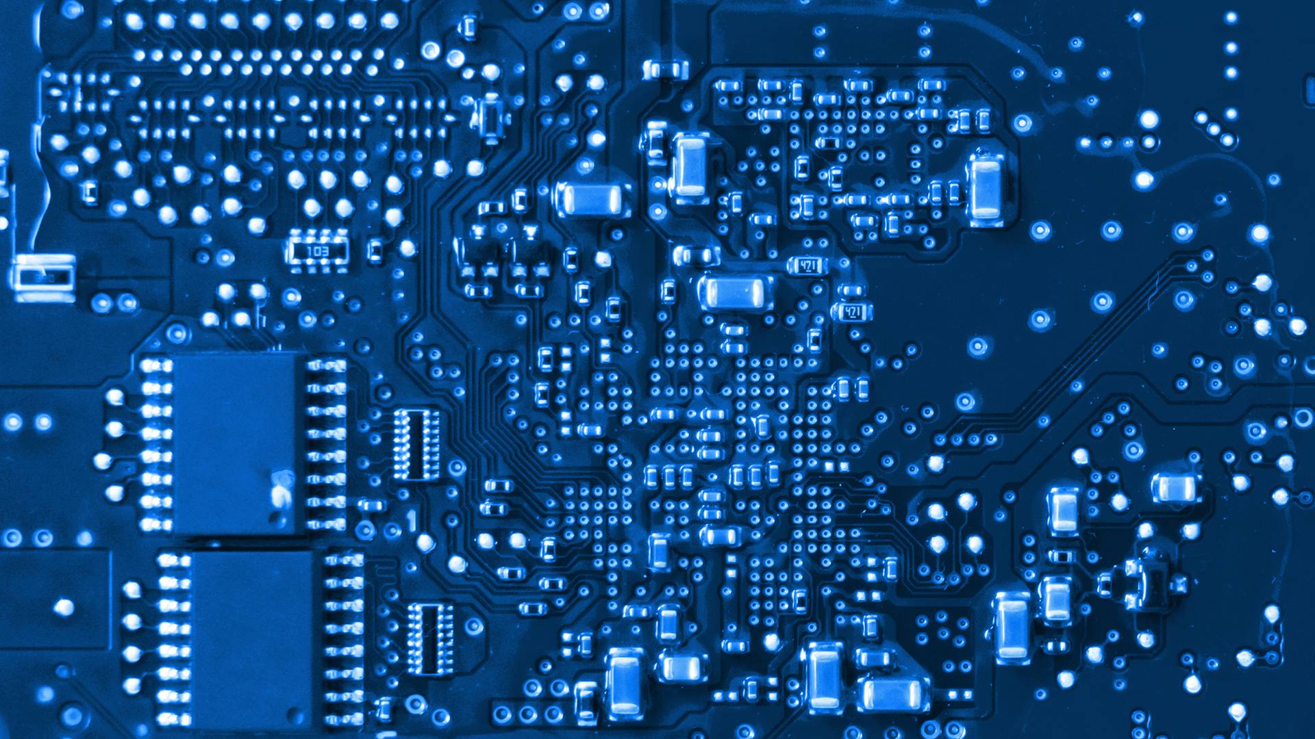 FGV Impianti Biella S.R.L. Circuito stampato con microchip e piste elettroniche FGV Impianti Biella S.R.L. Primo piano di una scheda elettronica blu ricoperta da fitte piste conduttive chiare. Sono presenti vari microchip rettangolari neri e componenti elettronici SMD disposti con precisione. Le linee di collegamento formano una rete complessa che occupa tutta la superficie visibile.