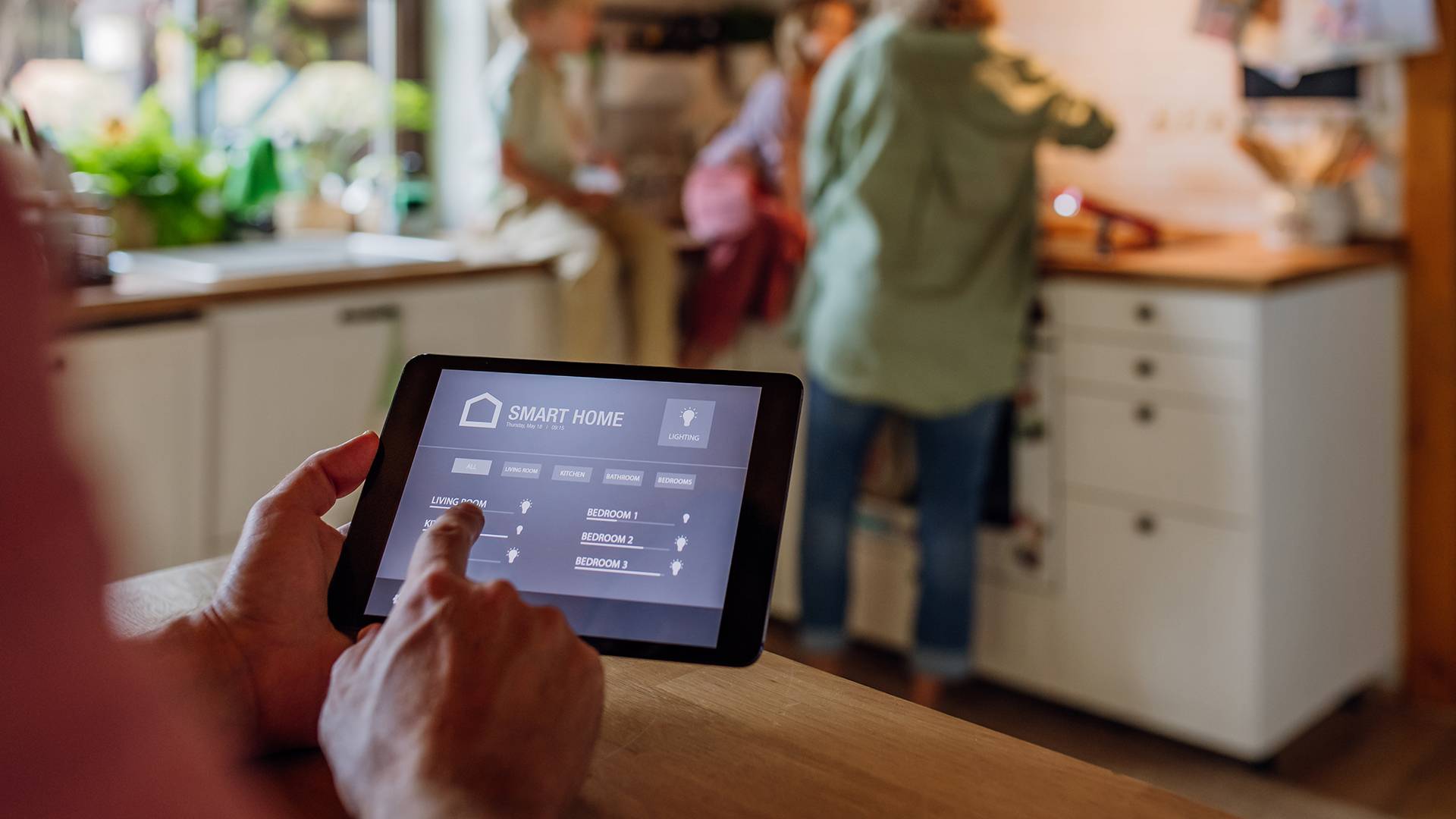 Persona che usa tablet per controllare dispositivi smart in cucina FGV Impianti Biella S.R.L. Interno di una cucina abitata con mobili moderni e piano di lavoro in legno. In primo piano una persona tiene in mano un tablet con interfaccia a icone e menu di controllo visibili sullo schermo (domotica). Sullo sfondo sfocato ci sono un’altra persona con due bambini seduti sul piano cucina, con alcuni oggetti domestici, arredi chiari, piante e luce naturale proveniente da una finestra.