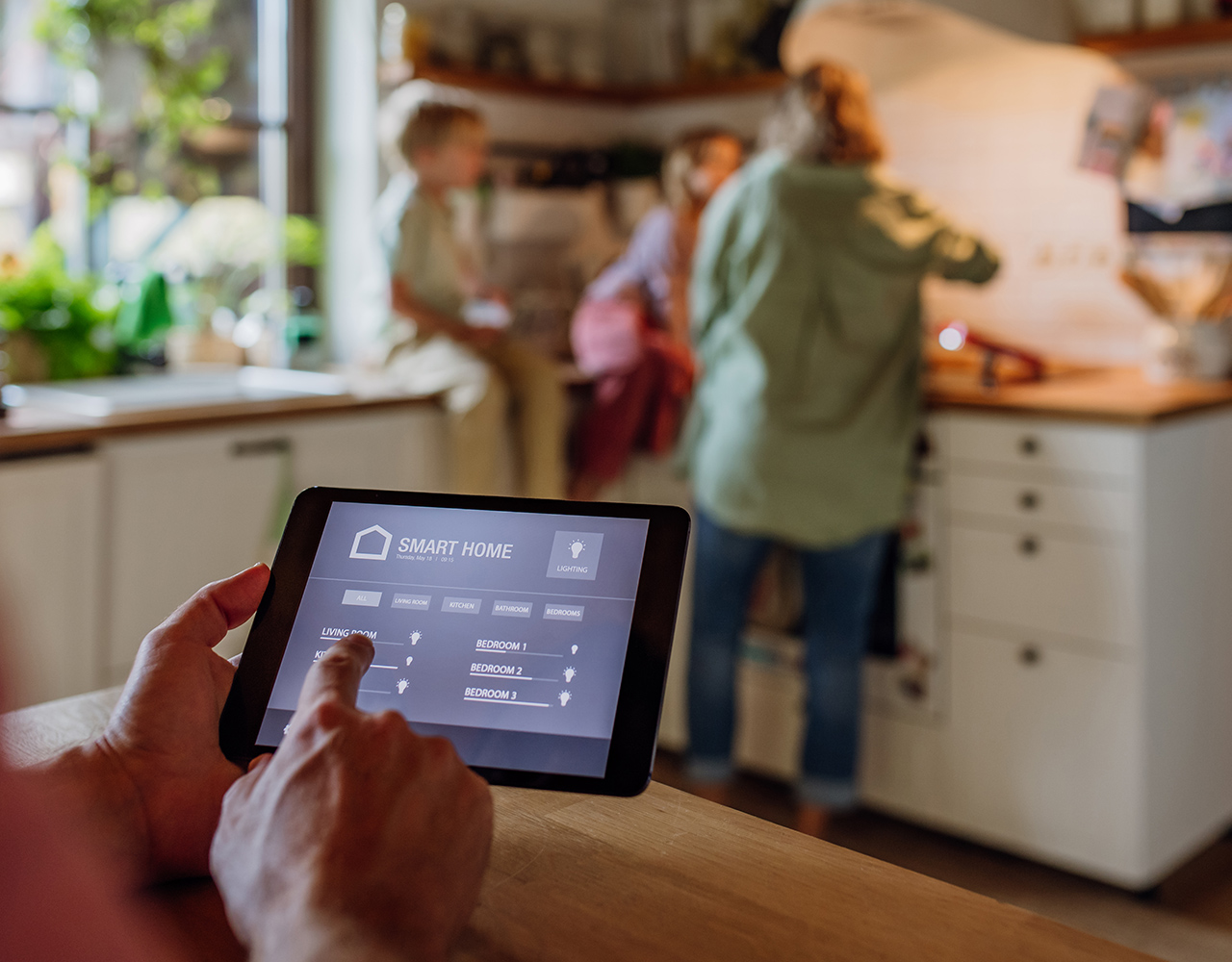 FGV Impianti Biella S.R.L. Persona che usa tablet per controllare dispositivi smart in cucina
Interno di una cucina abitata con mobili moderni e piano di lavoro in legno. In primo piano una persona tiene in mano un tablet con interfaccia a icone e menu di controllo visibili sullo schermo (domotica). Sullo sfondo sfocato ci sono un’altra persona con due bambini seduti  sul piano cucina, con alcuni oggetti domestici, arredi chiari, piante e luce naturale proveniente da una finestra.