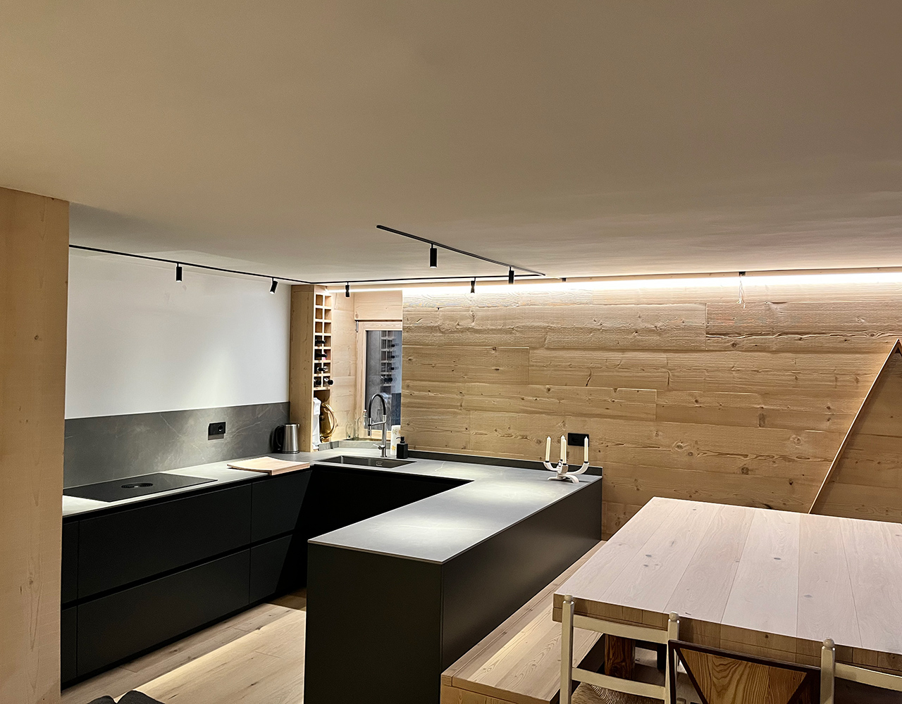 FGV Impianti Biella S.R.L. Cucina open space moderna con isola centrale e arredi in legno. Interno di una cucina ad induzione open space dal design contemporaneo. Da un lato troviamo un atgliere di legno, un bollitore in acciaio, un mobile porta bottiglie, una finestra e il lavandino. Al centro si trova un’isola scura con piano di lavoro su cui è appoggiato un candelabro moderno bianco a 4 bracci. e un tavolo in legno chiaro. Le pareti sono rivestite in pannelli lignei e i mobili hanno finiture nere opache. Il soffitto presenta una striscia luminosa a led che corre lungo il bordo e un binario con faretti orientabili. Sul lato destro pensile a forma triangolare