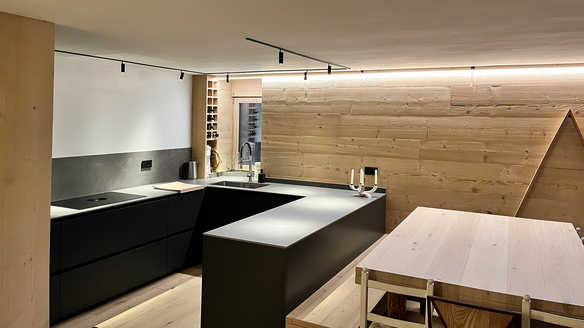 FGV Impianti Biella S.R.L. Cucina open space moderna con isola centrale e arredi in legno. Interno di una cucina ad induzione open space dal design contemporaneo. Da un lato troviamo un atgliere di legno, un bollitore in acciaio, un mobile porta bottiglie, una finestra e il lavandino. Al centro si trova un’isola scura con piano di lavoro su cui è appoggiato un candelabro moderno bianco a 4 bracci. e un tavolo in legno chiaro. Le pareti sono rivestite in pannelli lignei e i mobili hanno finiture nere opache. Il soffitto presenta una striscia luminosa a led che corre lungo il bordo e un binario con faretti orientabili. Sul lato destro pensile a forma triangolare