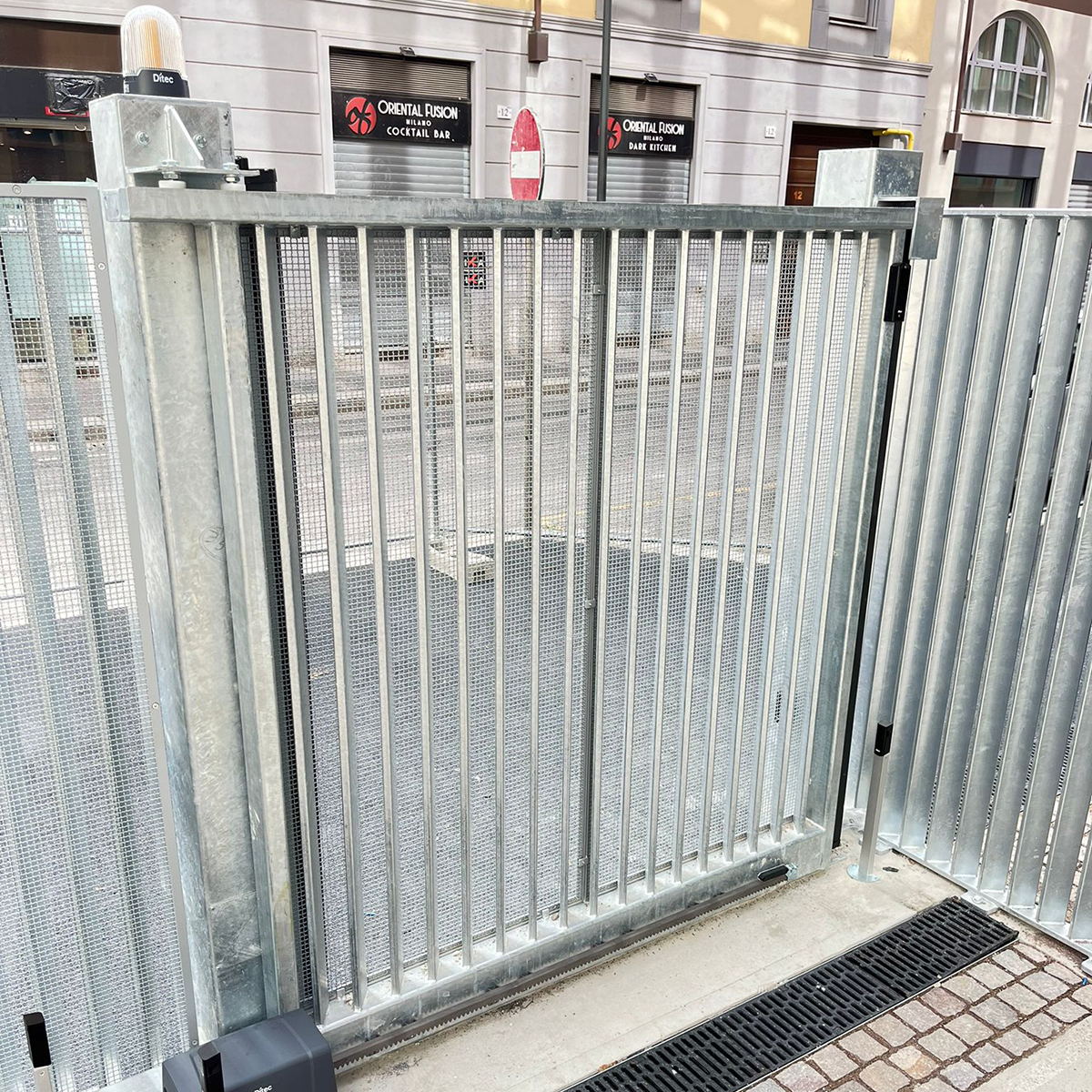 Fgv Impianti Biella Srl Automazioni Cancello scorrevole metallico con motore elettrico, strada, cartello e serrande dei negozi
Cancello scorrevole in metallo zincato composto da pannelli verticali e montato su una guida a terra fra due pilastri. Alla base è visibile l’alloggiamento del motore elettrico per l’automazione, con scatola di comando incassata e cablaggio protetto. Davanti al cancello si estende una porzione di marciapiede con un cartello di divieto di accesso e la carreggiata, su cui si affacciano vetrine e serrande metalliche dei negozi, alcune abbassate.