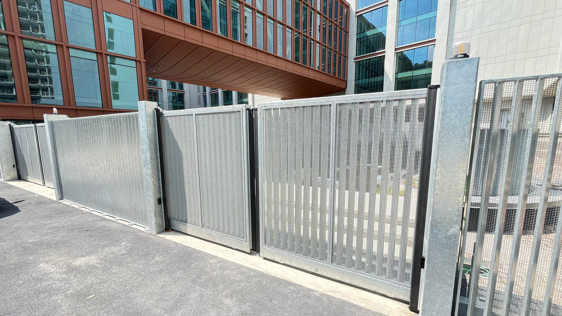 FGV Impianti Biella S.R.L. Cancello scorrevole metallico automatizzato con paesaggio montano Cancello scorrevole in metallo zincato montato su un vialetto pavimentato in pietra. Alla base, sul lato destro, è visibile il motore per l’apertura automatizzata. Il cancello è affiancato da colonne e ringhiere dello stesso materiale. Sullo sfondo si trovano un prato, diversi alberi verdi, case sparse e montagne coperte di vegetazione, sotto un cielo azzurro con nuvole bianche.