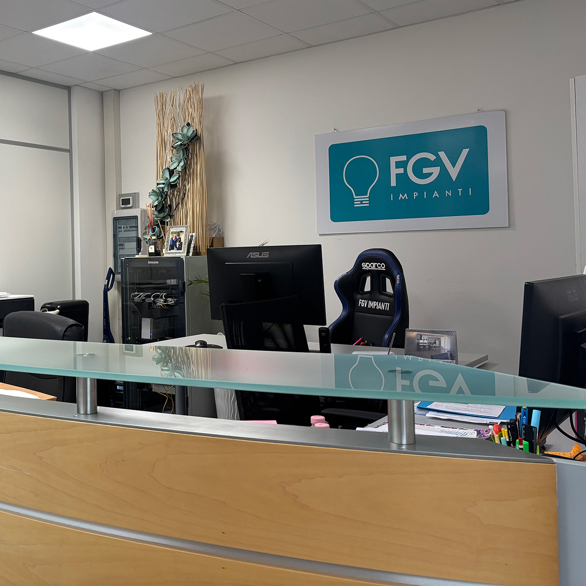 Reception aziendale con bancone in legno chiaro e logo FGV Impianti Biella Srl
Ambiente interno aziendale con bancone reception curvo in legno chiaro e superficie in vetro. Sul fondo è appeso un pannello con il logo FGV Impaint Biella Srl e lampade a soffitto illuminano uniformemente lo spazio. Si intravedono sedie da ufficio, monitor, arredi moderni e armadi elettrici.