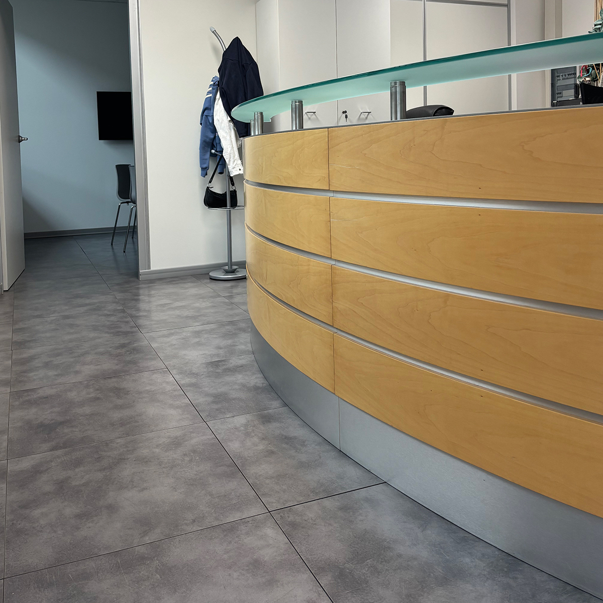 Reception aziendale con pavimento in gres e ingresso ufficio
Reception  aziendale di FGV impianti Biella srl con pavimento in gres grigio e pareti bianche. Sulla destra si vede parte del bancone reception in legno chiaro, mentre sul fondo c’è una porta d’ingresso aperta che conduce a un ufficio. Un appendiabiti con cappotti è posizionato accanto alla porta.