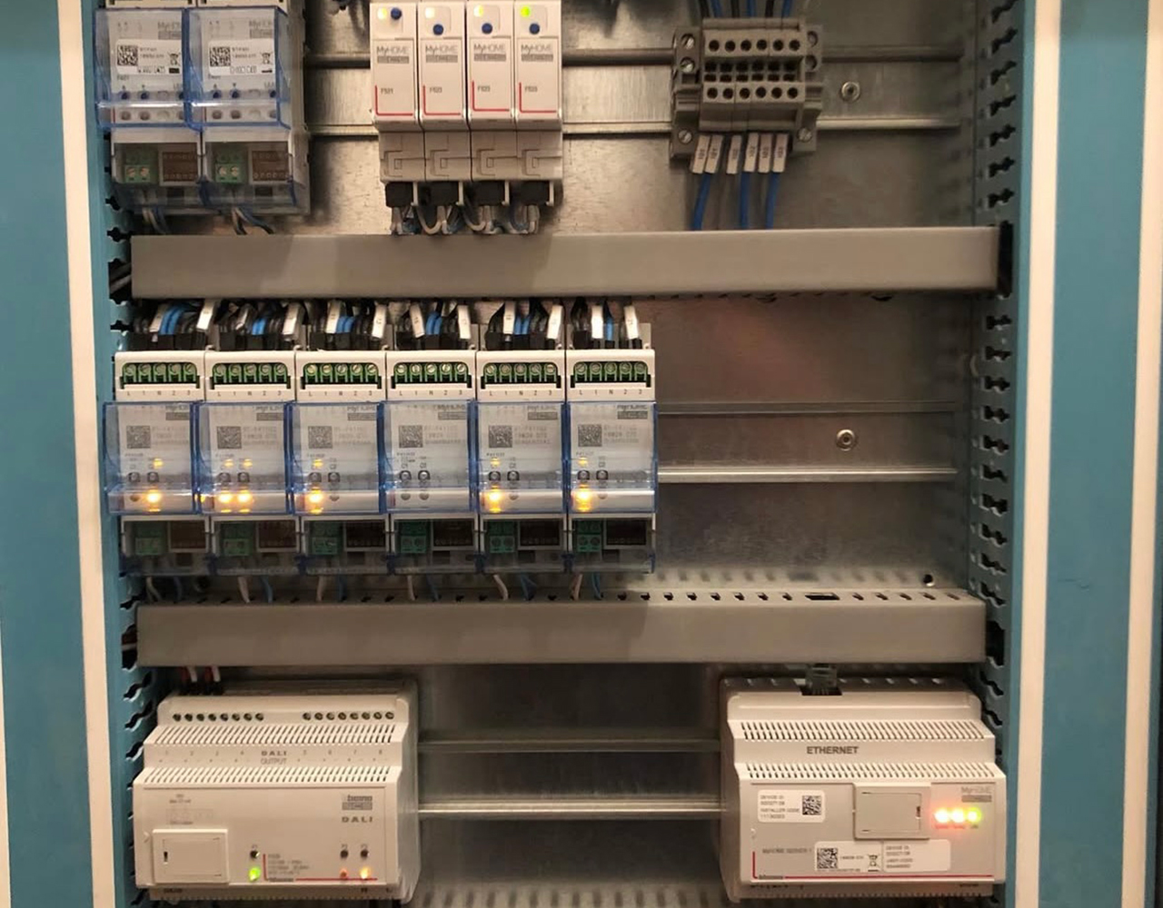FGV Impianti Biella S.R.L. Domotica Quadro elettrico industriale con interruttori e cablaggio ordinato.
Quadro elettrico montato a parete con interruttori magnetotermici, salvavita, relè e dispositivi di protezione organizzati su guide DIN. I cablaggi sono ordinati e raccolti in canaline grigie, con etichette identificative per una distribuzione sicura e precisa dell’energia.