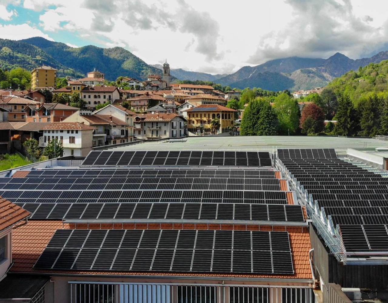 FGV Impianti Biella S.R.L. Pannelli solari su tetti di edifici con paesaggio montano Ampia superficie di pannelli fotovoltaici neri installati sui tetti rossi di un complesso industriale. In secondo piano si vedono altre case del paese circondate da alberi. Sullo sfondo si ergono montagne verdi e boscose. Il cielo sopra è parzialmente coperto da nuvole bianche e grigie, con tratti di azzurro visibile.