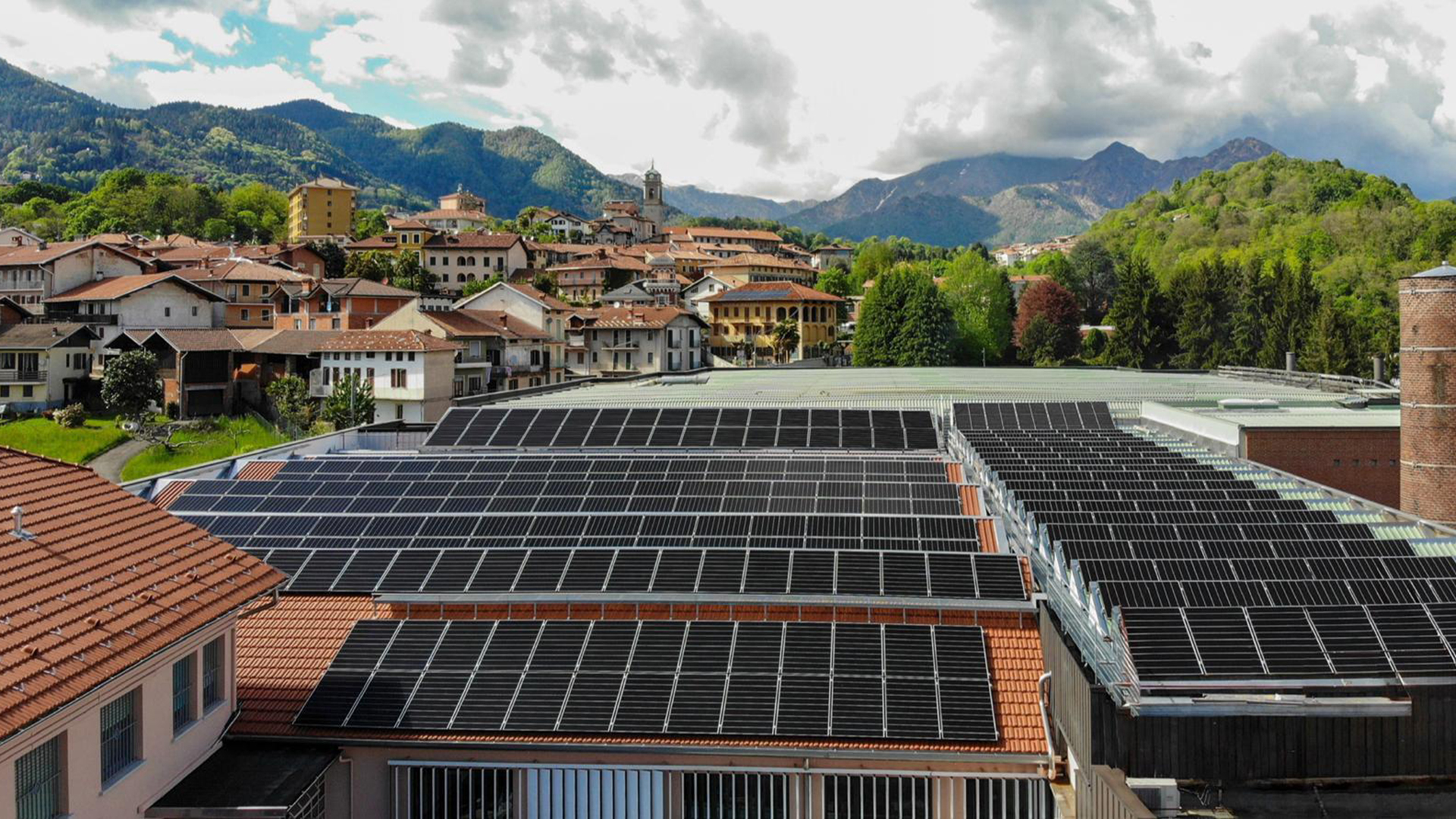 FGV Impianti Biella S.R.L. Pannelli solari su tetti di edifici con paesaggio montano Ampia superficie di pannelli fotovoltaici neri installati sui tetti rossi di un complesso industriale. In secondo piano si vedono altre case del paese circondate da alberi. Sullo sfondo si ergono montagne verdi e boscose. Il cielo sopra è parzialmente coperto da nuvole bianche e grigie, con tratti di azzurro visibile.