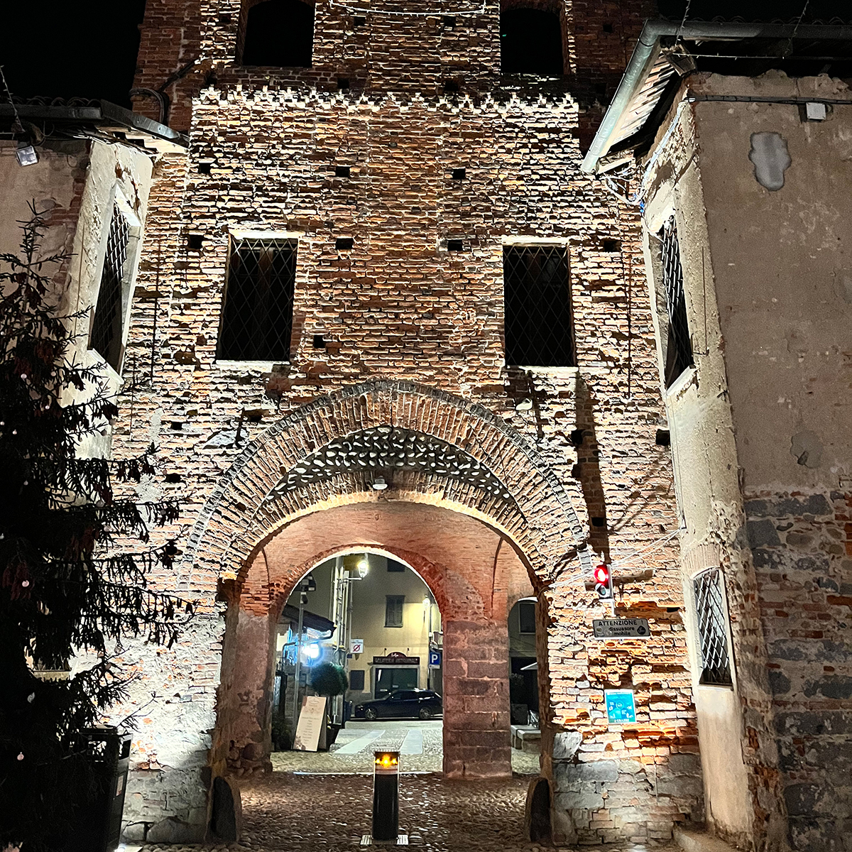 Fgv Impianti Biella Srl Impianti Civili Uscita Ricetto Candelo Arco in mattoni medievale con torre e ingresso Struttura architettonica dell'uscita del Ricetto di Candelo in mattoni rossi con grande arco centrale che funge da passaggio. Sopra l’arco si eleva una torre con finestre e muratura irregolare. L’interno dell’arco mostra un corridoio illuminato artificialmente. Alla sinistra si intravede un pino e al centro un dissuasore mobile con luce gialla nella sommità