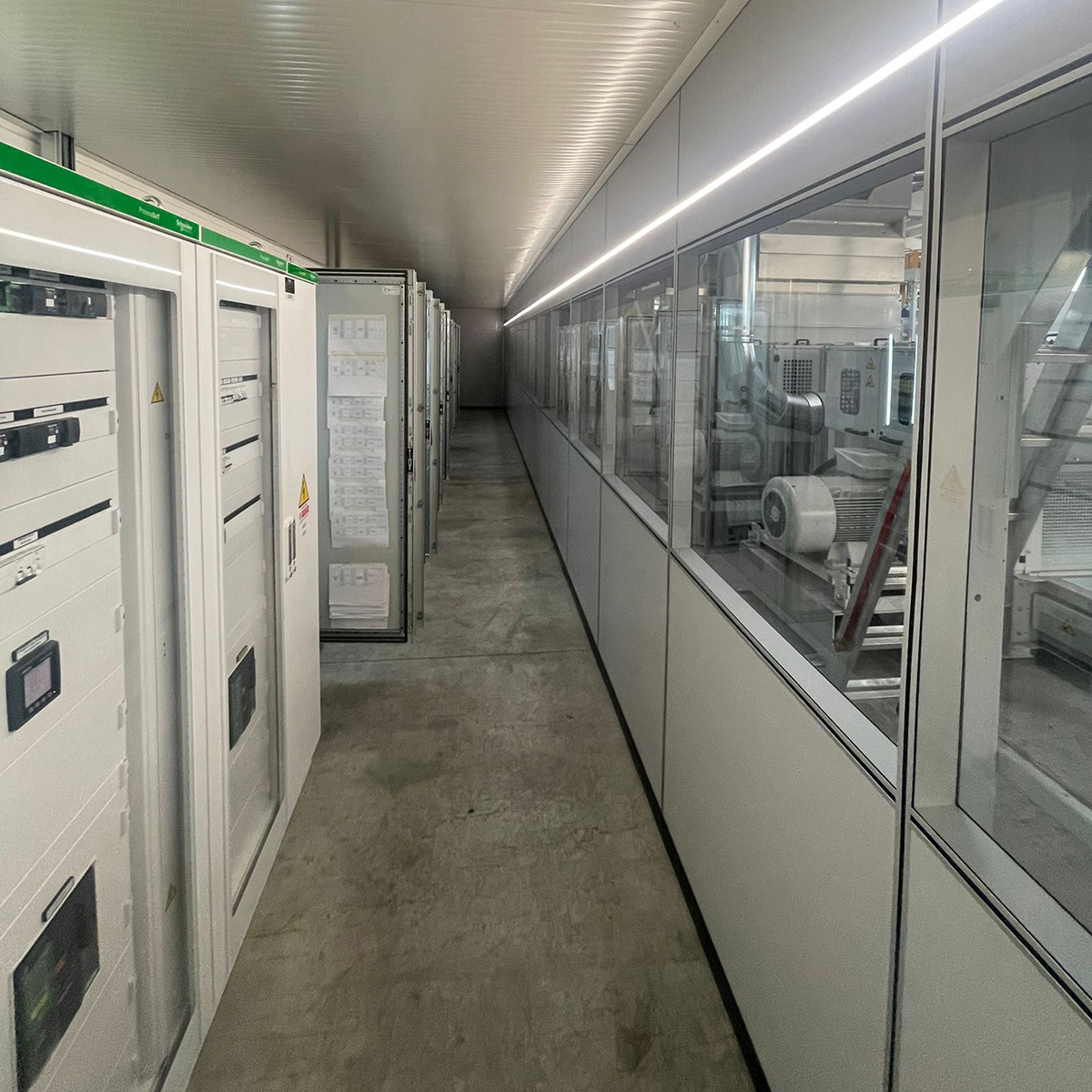FGV Impianti Biella S.R.L. Impianti Industriali Corridoio tecnico con quadri elettrici e illuminazione a neon
Descrizione: Lungo corridoio industriale con pavimento in cemento e pareti bianche. Su un lato sono installati grandi quadri elettrici con sportelli e indicatori luminosi, dall'altro lato ampie vetrate si affacciano sulla zona produttiva. Il soffitto è dotato di una fila di luci al neon che illuminano uniformemente lo spazio.