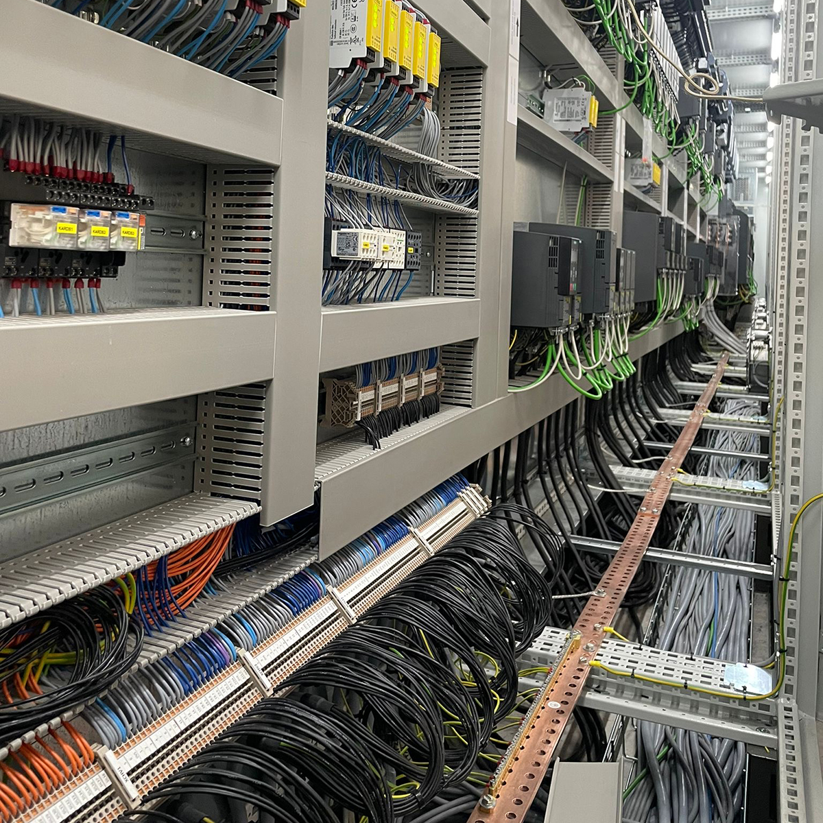 FGV Impianti Biella S.R.L. Impianti Industriali Quadro elettrico industriale con dispositivi e cablaggi ordinati Armadio elettrico industriale visto in prospettiva. Al suo interno ci sono file di dispositivi elettronici di vari colori, moduli di controllo, PLC e relè montati su rack. I cavi scorrono ordinati nelle canaline, con etichette di identificazione. Il corridoio tecnico si estende in profondità mostrando altri quadri allineati lungo la parete.