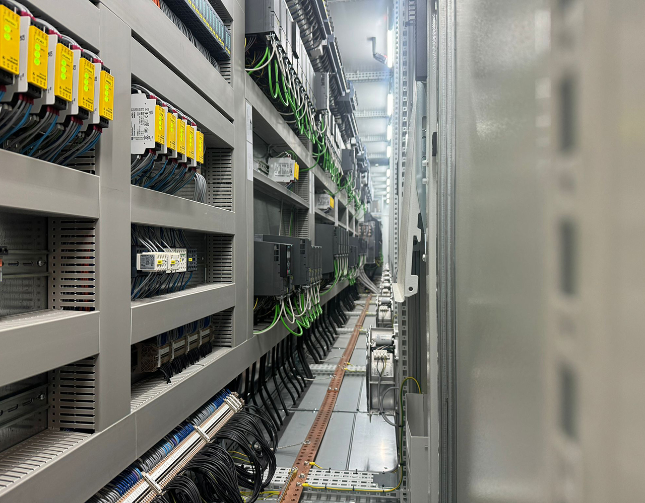 FGV Impianti Biella S.R.L. Quadro elettrico industriale con dispositivi e cablaggi ordinati Armadio elettrico industriale visto in prospettiva. Al suo interno ci sono file di dispositivi elettronici di vari colori, moduli di controllo, PLC e relè montati su rack. I cavi scorrono ordinati nelle canaline, con etichette di identificazione. Il corridoio tecnico si estende in profondità mostrando altri quadri allineati lungo la parete.