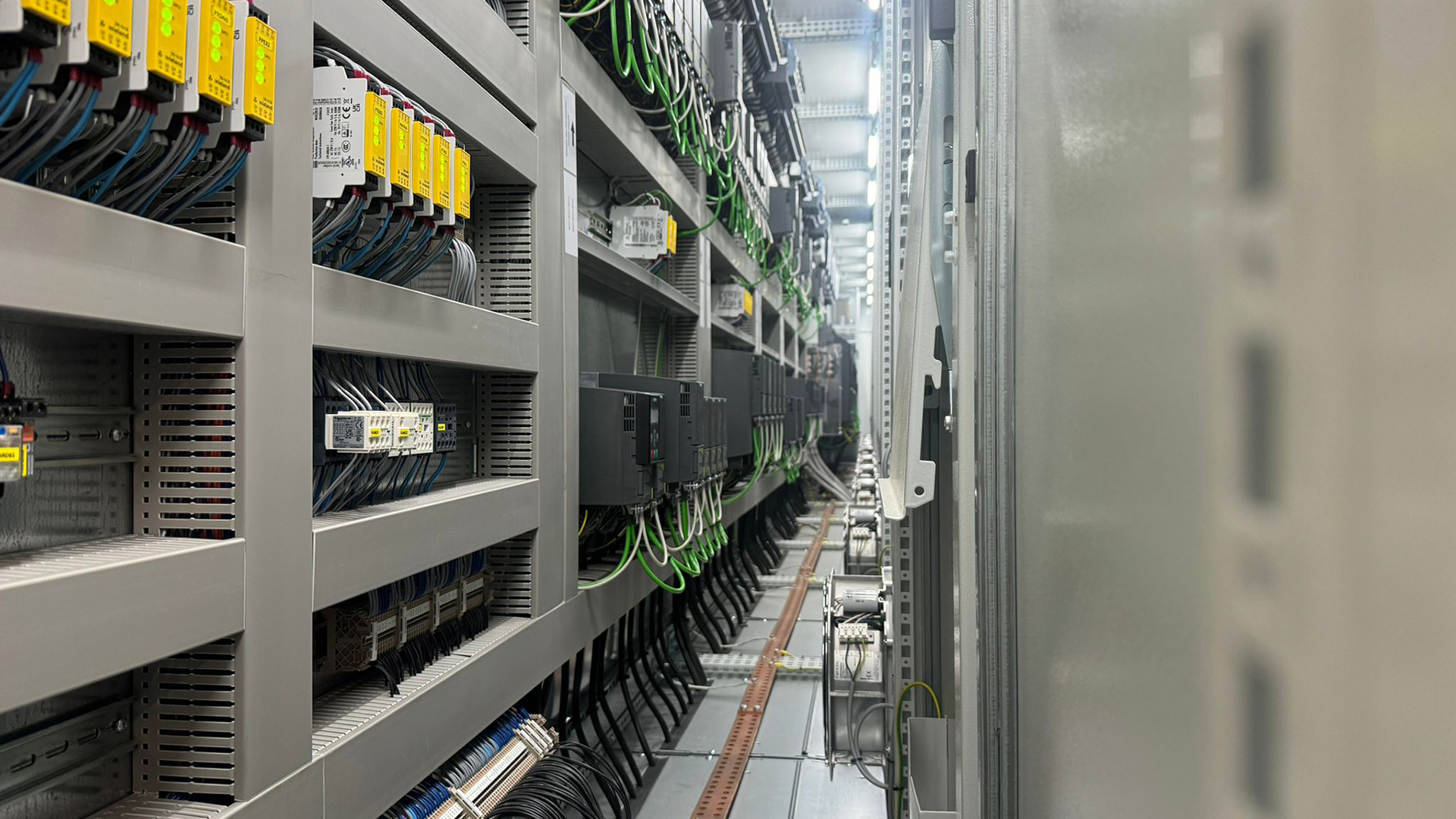 FGV Impianti Biella S.R.L. Quadro elettrico industriale con dispositivi e cablaggi ordinati Armadio elettrico industriale visto in prospettiva. Al suo interno ci sono file di dispositivi elettronici di vari colori, moduli di controllo, PLC e relè montati su rack. I cavi scorrono ordinati nelle canaline, con etichette di identificazione. Il corridoio tecnico si estende in profondità mostrando altri quadri allineati lungo la parete.