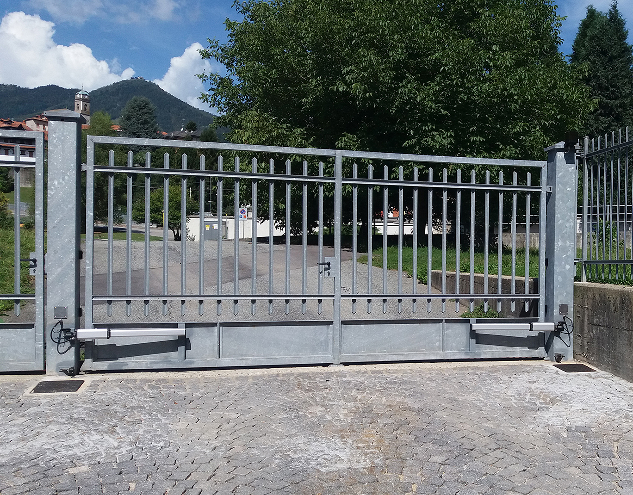 FGV Impianti Biella S.R.L. Cancello scorrevole metallico automatizzato con paesaggio montano Cancello scorrevole in metallo zincato montato su un vialetto pavimentato in pietra. Alla base, sul lato destro, è visibile il motore per l’apertura automatizzata. Il cancello è affiancato da colonne e ringhiere dello stesso materiale. Sullo sfondo si trovano un prato, diversi alberi verdi, case sparse e montagne coperte di vegetazione, sotto un cielo azzurro con nuvole bianche.