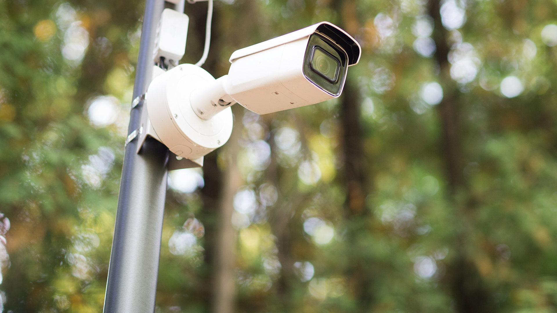 FGV Impianti Biella S.R.L. Telecamera di sicurezza bianca montata su supporto esterno Telecamera di sorveglianza bianca di tipo bullet, fissata a un supporto metallico esterno. L’obiettivo è orientato frontalmente. Sullo sfondo si distinguono alberi verdi sfocati illuminati dalla luce naturale del giorno.