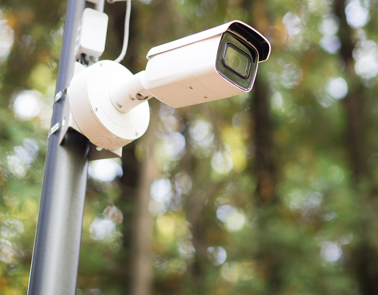 FGV Impianti Biella S.R.L. Telecamera di sicurezza bianca montata su supporto esterno Telecamera di sorveglianza bianca di tipo bullet, fissata a un supporto metallico esterno. L’obiettivo è orientato frontalmente. Sullo sfondo si distinguono alberi verdi sfocati illuminati dalla luce naturale del giorno.