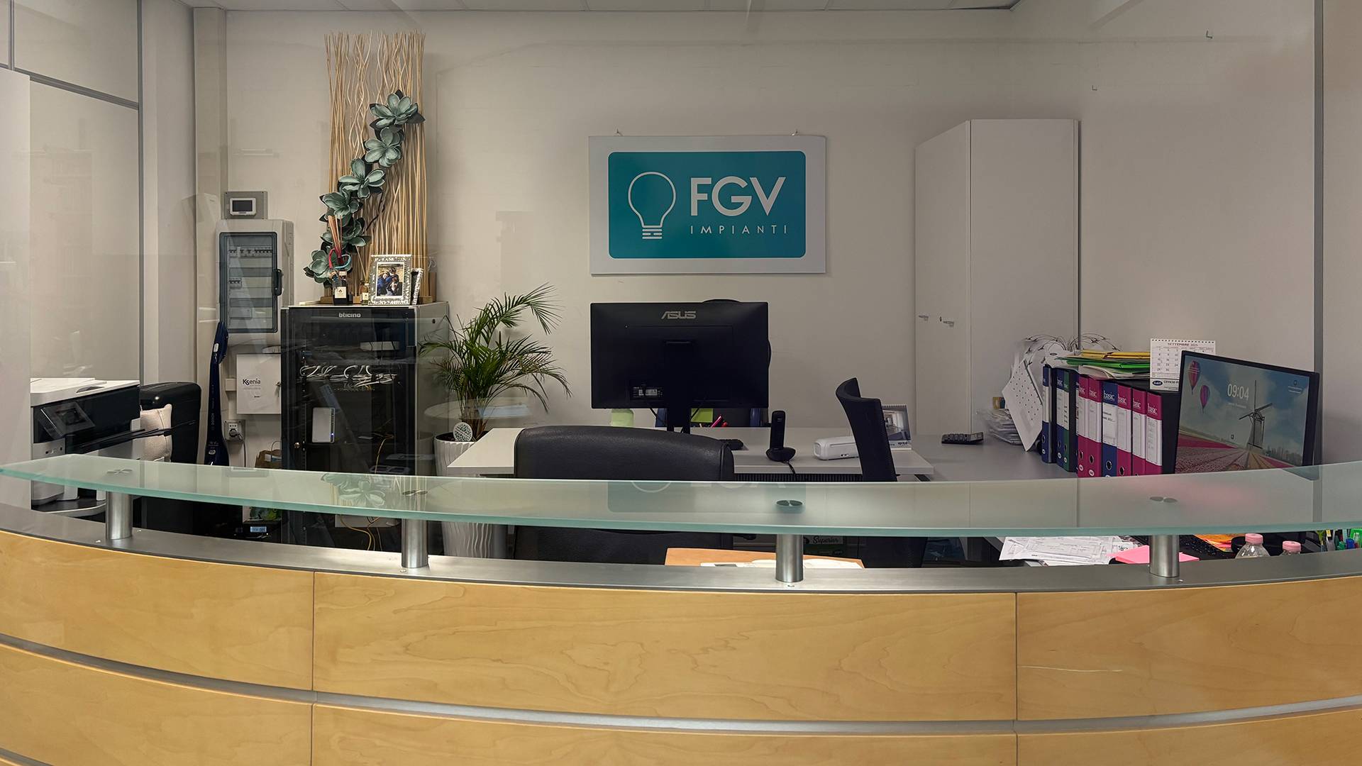 Reception aziendale di FGV impianti Biella Srl con bancone curvo e postazioni operative Vista frontale della reception aziendale con bancone in legno chiaro e vetro superiore. Dietro si trovano due postazioni operative con computer, monitor, stampanti e raccoglitori colorati. Sul muro è appeso il logo FGV impianti Biella srl e in un angolo si nota una pianta decorativa e armadi elettrici