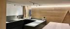 FGV Impianti Biella S.R.L. Cucina open space moderna con isola centrale e arredi in legno. Interno di una cucina ad induzione open space dal design contemporaneo. Da un lato troviamo un atgliere di legno, un bollitore in acciaio, un mobile porta bottiglie, una finestra e il lavandino. Al centro si trova un’isola scura con piano di lavoro su cui è appoggiato un candelabro moderno bianco a 4 bracci.  e un tavolo in legno chiaro. Le pareti sono rivestite in pannelli lignei e i mobili hanno finiture nere opache. Il soffitto presenta una striscia luminosa a led che corre lungo il bordo e un binario con faretti orientabili. Sul lato destro pensile a forma triangolare