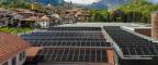FGV Impianti Biella S.R.L. Pannelli solari su tetti di edifici con paesaggio montano Ampia superficie di pannelli fotovoltaici neri installati sui tetti rossi di un complesso industriale. In secondo piano si vedono altre case del paese circondate da alberi. Sullo sfondo si ergono montagne verdi e boscose. Il cielo sopra è parzialmente coperto da nuvole bianche e grigie, con tratti di azzurro visibile.